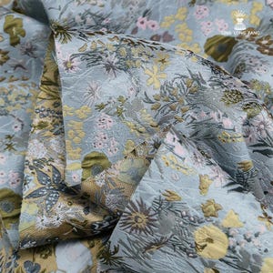 May include: A light blue fabric featuring a woven floral pattern in shades of yellow, olive green, and soft pink. The textile displays a textured surface with intricate botanical details and the text LING LONG FANG printed in the upper right corner.