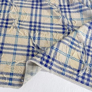 May include: A fabric featuring a cream-colored base with a blue plaid pattern. The material displays a textured, frayed effect along the lines of the check design, creating a dimensional appearance across the surface of the textile.