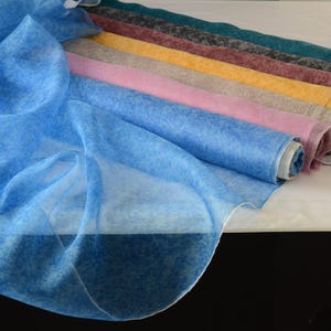 Può includere: Una selezione di rotoli di tessuto colorati, tra cui tonalità di blu, rosa, giallo e grigio. Il tessuto ha un aspetto delicato e trasparente, con un sottile motivo maculato. I rotoli sono disposti in una fila diagonale, mostrando la varietà di colori e trame.