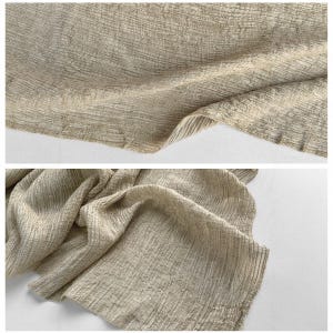 May include: Close-up of a textured, beige fabric with a crinkled appearance. The fabric has a subtle sheen and a woven pattern, creating a soft, flowing drape. The image shows the fabric in two different arrangements, highlighting its texture and movement.