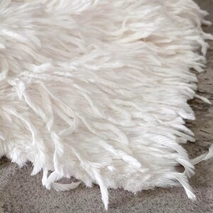 Puede incluir: Una alfombra de piel sintética blanca con fibras largas, peludas y tenues que crean una apariencia texturizada y en capas. El material tiene un tono blanco suave con hebras individuales que están ligeramente rizadas y fluidas.