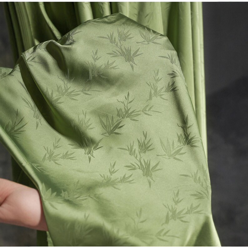 May include: Close-up of a light green fabric with a subtle bamboo leaf pattern. The fabric has a smooth, silky texture and a slight sheen. The folds of the fabric are visible, suggesting it is part of a garment or drapery.