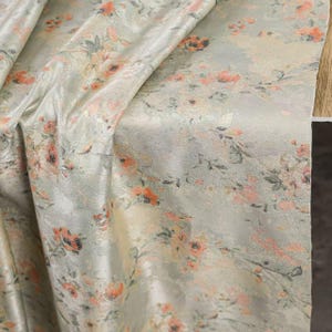 May include: A draped fabric with a floral pattern. The fabric has a light gray background with a subtle sheen. The floral design features orange and pink flowers with green leaves, creating a soft, vintage aesthetic. The fabric is folded, showing its texture and drape.