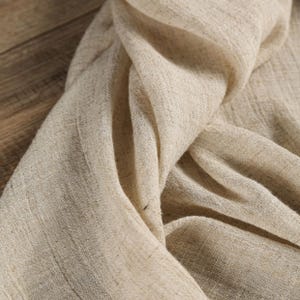 May include: Close-up of a beige linen fabric with a textured weave. The fabric is draped, creating soft folds and shadows. The neutral color and natural texture suggest a focus on organic materials and minimalist design.