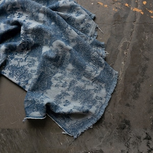 Könnte beinhalten: Blau-weiß gemusterter Stoff mit Landschaftsdesign. Der Stoff ist auf einer dunklen Oberfläche drapiert und zeigt einen detaillierten Druck von Bäumen, Gebäuden und anderen natürlichen Elementen. Die Kanten sind ausgefranst.