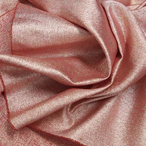 May include: A piece of fabric in a dusty rose pink color with a metallic gold shimmer finish. The material is draped in soft folds, highlighting the reflective quality of the surface and the fine, granular texture of the gold-toned coating.