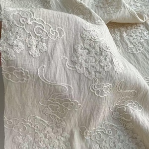 Puede incluir: Tela blanquecina con un patrón de nubes bordadas. La tela es suave y fluida, con costuras blancas detalladas que crean un diseño repetitivo de nubes estilizadas. La textura de la tela es visible.