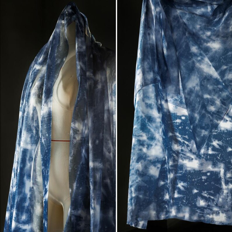May include: A sheer fabric with a blue and white tie-dye pattern. The fabric is draped over a mannequin, highlighting its flow and texture. The design resembles clouds or water, creating a soft, airy aesthetic. The fabric is suitable for clothing.
