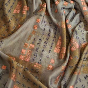 May include: Close-up of a draped fabric with a blend of gray and brown tones. The fabric features copper-colored square accents and black calligraphic script. The overall texture is soft and flowing, suggesting a scarf or shawl.
