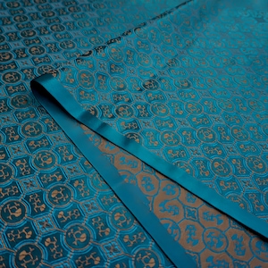 Może przedstawiać: Zbliżenie turkusowej tkaniny z powtarzającym się wzorem złotych kwiatowych i geometrycznych kształtów. Tkanina ma satynowy połysk i szczegół w postaci złożonej wstążki. Ogólny design jest bogaty i luksusowy, odpowiedni do odzieży lub wystroju domu.