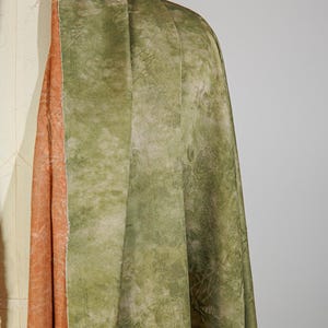 May include: Close-up of a draped fabric with a textured, mottled appearance. The outer layer is a muted green with darker, organic patterns. The fabric's interior reveals a contrasting peach-colored layer, adding depth and visual interest to the textile.