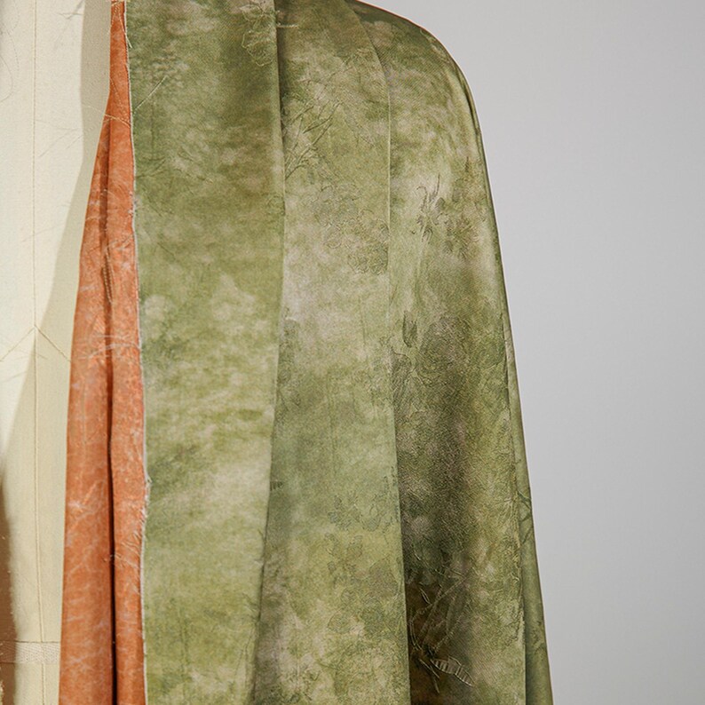 May include: Close-up of a draped fabric with a textured, mottled appearance. The outer layer is a muted green with darker, organic patterns. The fabric's interior reveals a contrasting peach-colored layer, adding depth and visual interest to the textile.