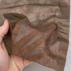 Puede incluir: Primer plano de un trozo de tela marrón translúcida. La tela se sostiene, revelando su textura y la forma en que la luz la atraviesa. El material parece delicado y ligeramente arrugado.