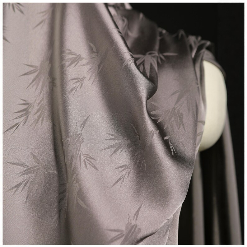 May include: Close-up of a draped, silvery-gray fabric with a subtle bamboo leaf pattern. The fabric has a smooth, silky texture and is draped over a mannequin, showcasing its flow and drape. The background is dark.