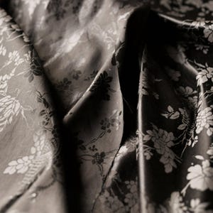May include: Close-up of a dark fabric with a floral pattern. The fabric is folded, creating shadows and highlights. The flowers are in a lighter shade, contrasting with the darker background. The overall effect is elegant and textured.