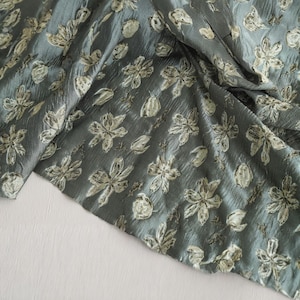 May include: A close-up of a teal fabric with a floral pattern. The fabric has a crinkled texture and is adorned with a repeating design of stylized flowers and buds in shades of cream and light gray.