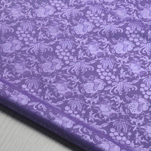 May include: A close-up of a lavender fabric with a repeating pattern of stylized fruit, flowers, and vines. The design is in a lighter shade of lavender, creating a subtle contrast. The fabric is folded, showing its texture and sheen.