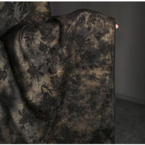 以下が含まれることがあります： 花柄の暗い生地のクローズアップ。生地は、金と黒の背景に黒い花柄のデザインが施されています。花柄にはバラと葉が含まれています。生地はドレープされており、その質感と流れを示しています。