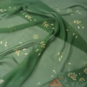 Puede incluir: Una tela fluida y transparente en tonos verdes, adornada con delicados diseños florales y de hojas amarillas y blancas. La tela está drapeada, mostrando su textura ligera y aireada, ideal para bufandas o prendas.