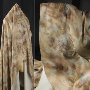 Könnte beinhalten: Ein drapierter Stoff mit einem gesprenkelten Muster in den Farbtönen Beige, Braun und einem Hauch von Blau. Der Stoff ist über einer Form drapiert, wodurch Falten und Schatten entstehen. Die Nahaufnahme zeigt die Textur und Farbvariationen.