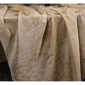May include: A draped, textured fabric in shades of beige and brown. The fabric has a subtle, repeating pattern of thin, white, vein-like lines. The material is folded and draped, creating shadows and depth. The overall appearance is rustic and natural.