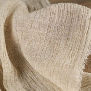 May include: Close-up of a beige linen fabric with a textured, wrinkled appearance. The fabric has a natural, organic look with visible fibers and a slightly frayed edge. The folds and shadows create depth.