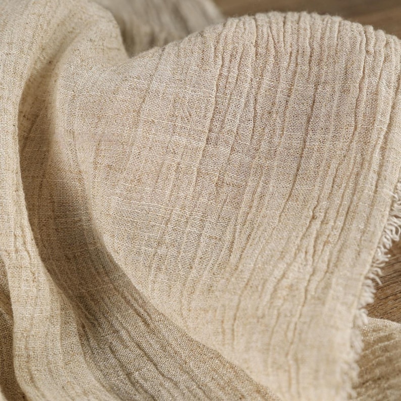 May include: Close-up of a beige linen fabric with a textured, wrinkled appearance. The fabric has a natural, organic look with visible fibers and a slightly frayed edge. The folds and shadows create depth.