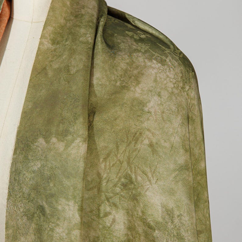 May include: Close-up of a green fabric with a subtle, textured pattern. The fabric drapes over a mannequin, showcasing its flow and design. The color palette includes shades of green and beige, creating a natural, earthy aesthetic. The fabric appears to be lightweight.