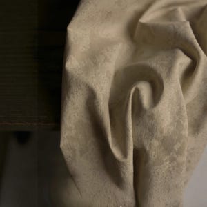 May include: Close-up of a beige fabric with a subtle, textured pattern. The fabric is draped, showing folds and shadows. The color is a neutral tone, suitable for various design applications.