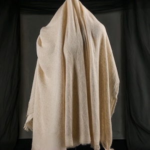 May include: A draped, off-white woven textile with a textured surface. The fabric is loosely woven, with visible variations in the weave. The edges of the textile are fringed. The background is a dark gray.