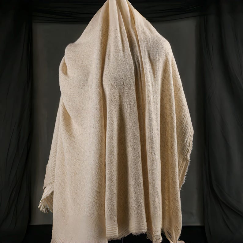 May include: A draped, off-white woven textile with a textured surface. The fabric is loosely woven, with visible variations in the weave. The edges of the textile are fringed. The background is a dark gray.