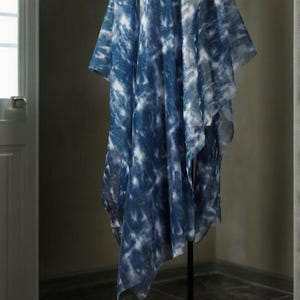 May include: A blue and white tie-dye patterned garment. The fabric is lightweight and has a loose, draped shape. The garment is displayed on a black stand, highlighting its design and texture. The garment's measurements are in centimeters.