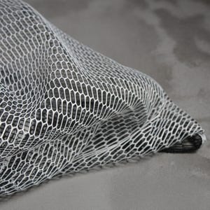 Puede incluir: Primer plano de una tela de malla blanca con un patrón de panal. La tela está drapeada, creando pliegues y sombras suaves. El fondo es gris sólido, proporcionando contraste con el intrincado diseño de la malla.