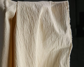 Può includere: Primo piano di un tessuto color crema con una superficie strutturata e stropicciata. Il tessuto è drappeggiato, rivelando il suo spessore e i bordi grezzi. Il materiale sembra essere lino o una fibra naturale simile, adatta all'abbigliamento o all'arredamento.