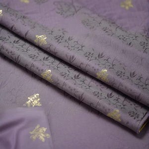 May include: Close-up of a folded fabric with a lavender hue. The fabric features a repeating pattern of gray floral designs and gold accents. The folds create shadows and depth, highlighting the texture and details of the material.