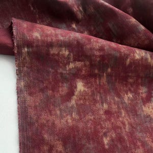 Puede incluir: Un trozo de tela con un patrón abstracto en burdeos y beige. La tela tiene una apariencia texturizada y un borde sin rematar. El patrón tiene un efecto acuarela, con áreas de beige claro y burdeos más oscuro.