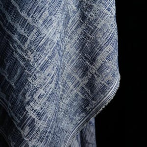 Op de afbeelding: Een stuk stof met een diepblauw en wit geweven patroon met een getextureerd, gearceerd uiterlijk. Het materiaal vertoont een zichtbare rasterachtige structuur met gerafelde randen aan de zijkant, wat een dicht, gelaagd textielontwerp laat zien.
