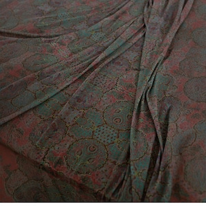 Op de afbeelding: Close-up van een stof met een paisley-patroon in tinten bordeauxrood, blauwgroen en bruin. De stof is gedrapeerd en toont plooien en textuur. Het patroon heeft cirkelvormige en bloemmotieven.