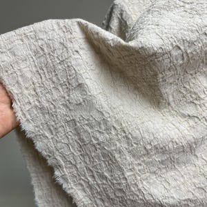 Könnte beinhalten: Ein Stück hellbeiger Stoff mit einer ausgeprägten, erhabenen und zerknitterten Textur, die ein gitterartiges Muster auf der Oberfläche erzeugt. Das Material weist an der Seite eine rohe, ausgefranste Kante auf, die die Webstruktur zeigt.