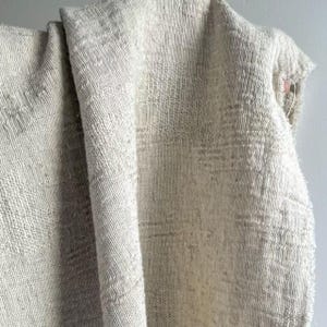 May include: A close-up of a textured, off-white fabric. The fabric has a woven appearance with a slightly irregular pattern. The material is draped, showing folds and shadows that highlight the texture. The overall look is neutral and natural.