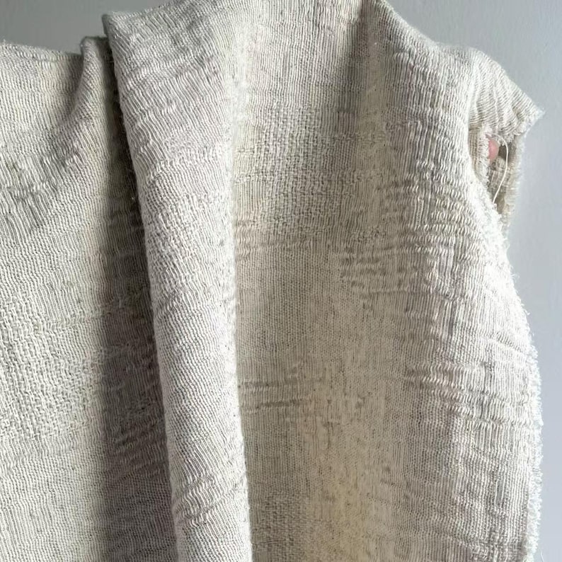 May include: A close-up of a textured, off-white fabric. The fabric has a woven appearance with a slightly irregular pattern. The material is draped, showing folds and shadows that highlight the texture. The overall look is neutral and natural.