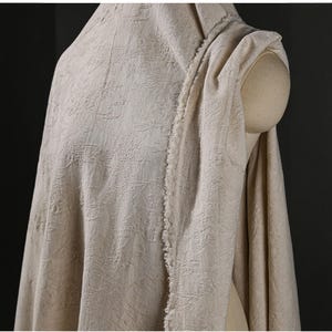 May include: Beige fabric draped over a mannequin, showcasing a detailed floral pattern. The fabric has a textured appearance and a raw edge, suggesting a natural fiber like linen or cotton. The neutral color and delicate design make it suitable for various textile projects.