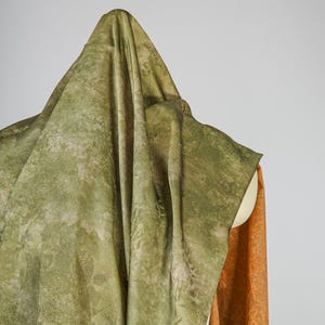 May include: A draped fabric in shades of green and tan, with a subtle botanical pattern. The fabric is draped over a form, with a glimpse of orange fabric visible on the side. The overall effect is soft and textured.
