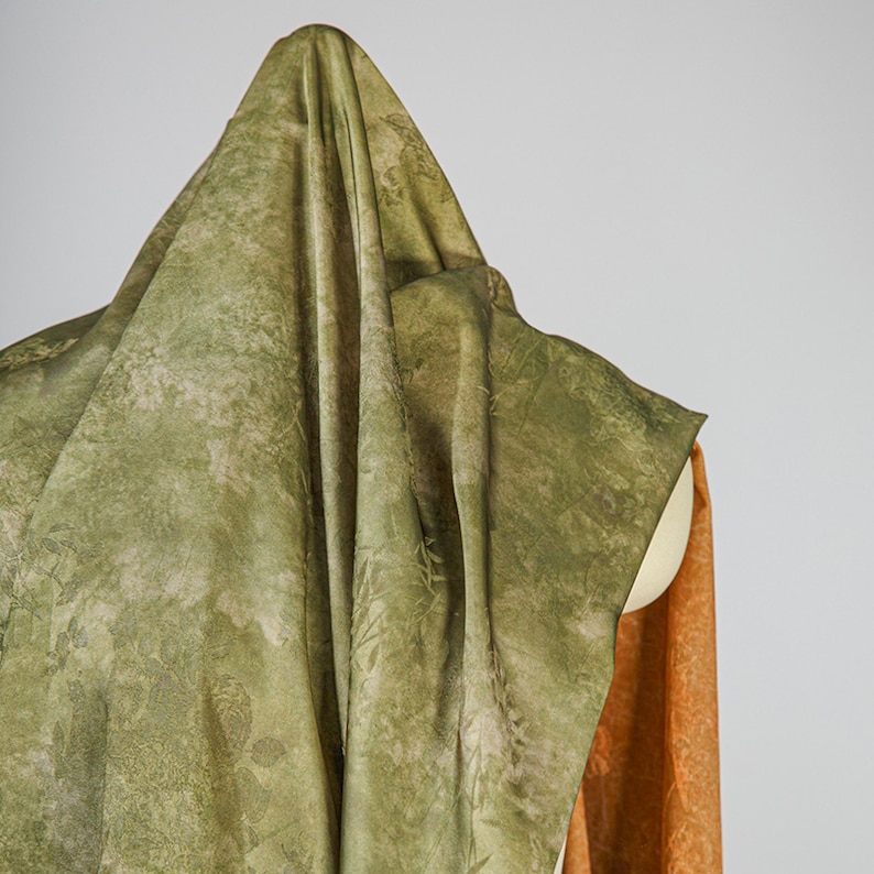 May include: A draped fabric in shades of green and tan, with a subtle botanical pattern. The fabric is draped over a form, with a glimpse of orange fabric visible on the side. The overall effect is soft and textured.