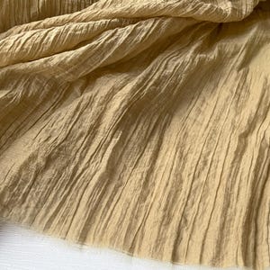 Pode incluir: Close-up de um tecido bege texturizado com pregas verticais. O tecido é drapeado, criando dobras e sombras que adicionam profundidade. O material parece macio e leve, adequado para roupas ou decoração de casa.