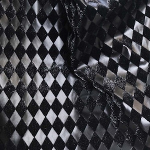 May include: Close-up of a fabric with a repeating diamond pattern. The diamonds alternate between black and silver, creating a textured, reflective surface. The fabric appears to be a shiny material, possibly satin or a similar textile.
