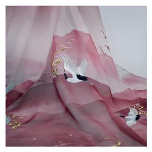 May include: Sheer fabric with a gradient of pink and burgundy hues, embroidered with white cranes, gold accents, and mountain silhouettes. The fabric is draped, creating soft folds and a flowing appearance.