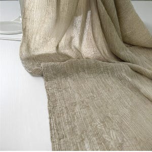 May include: A beige, textured fabric with a crinkled appearance. The fabric is draped over a white surface, with soft folds and subtle patterns visible throughout. The material appears lightweight and airy, suitable for scarves or decorative purposes.