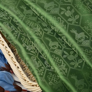 Puede incluir: Primer plano de una tela verde con un patrón repetido de ciervos, flores y formas geométricas. La tela está doblada, revelando su textura y brillo. Adecuada para tapicería o fines decorativos.