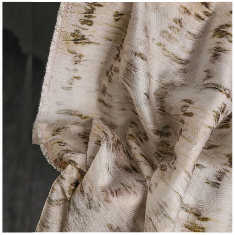 Puede incluir: Tela blanquecina con un patr&oacute;n texturizado con dise&ntilde;os en forma de pincelada marr&oacute;n y verde oliva. La tela est&aacute; drapeada, mostrando pliegues y un borde sin rematar, lo que sugiere un textil para proyectos de dise&ntilde;o.